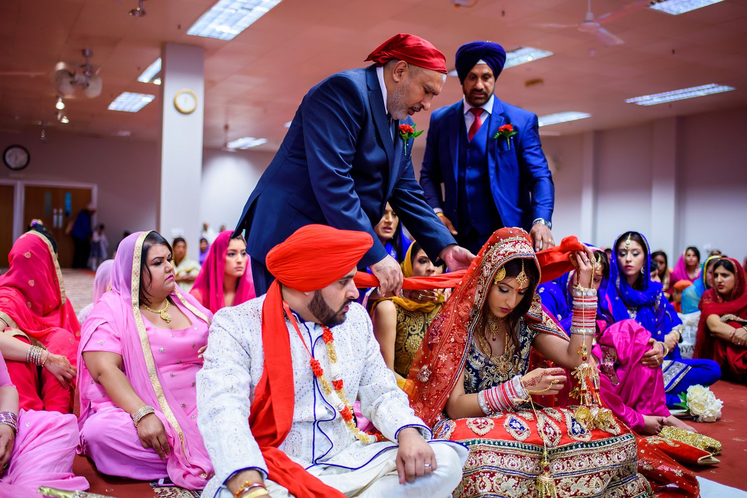 Group prayer ceremony during Sikh wedding with attendees seated on red carpet.
