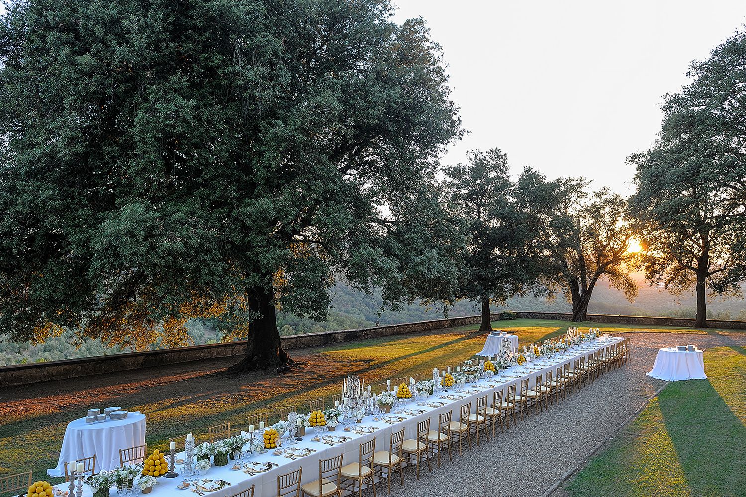 Long banquet table set for an elegant outdoor dinner party at sunset with white linens and tree-lined path.