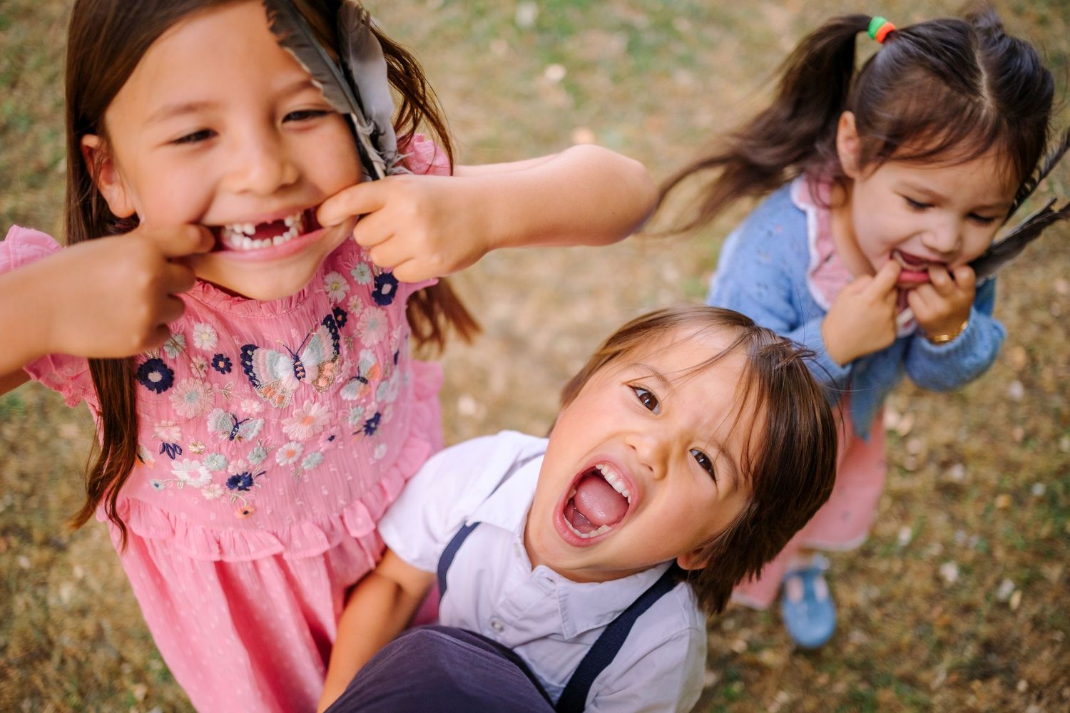 Three children playing and making silly faces outdoors while laughing together on a sunny day.