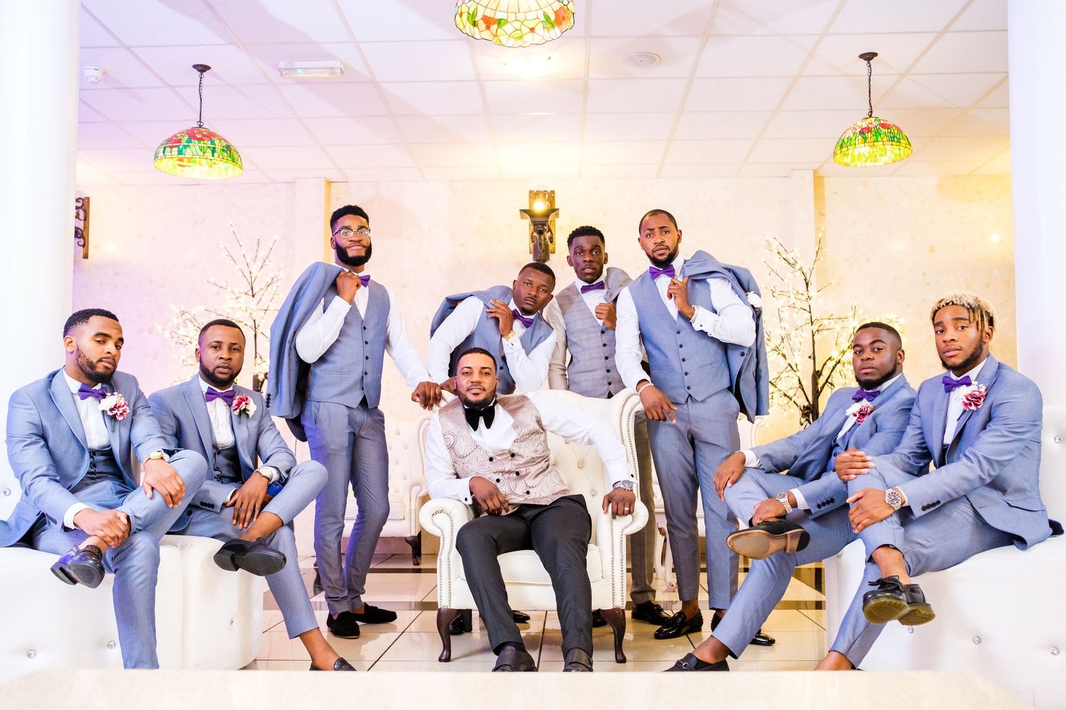 Groomsmen in light blue suits pose playfully for photos in an elegant white reception hall.