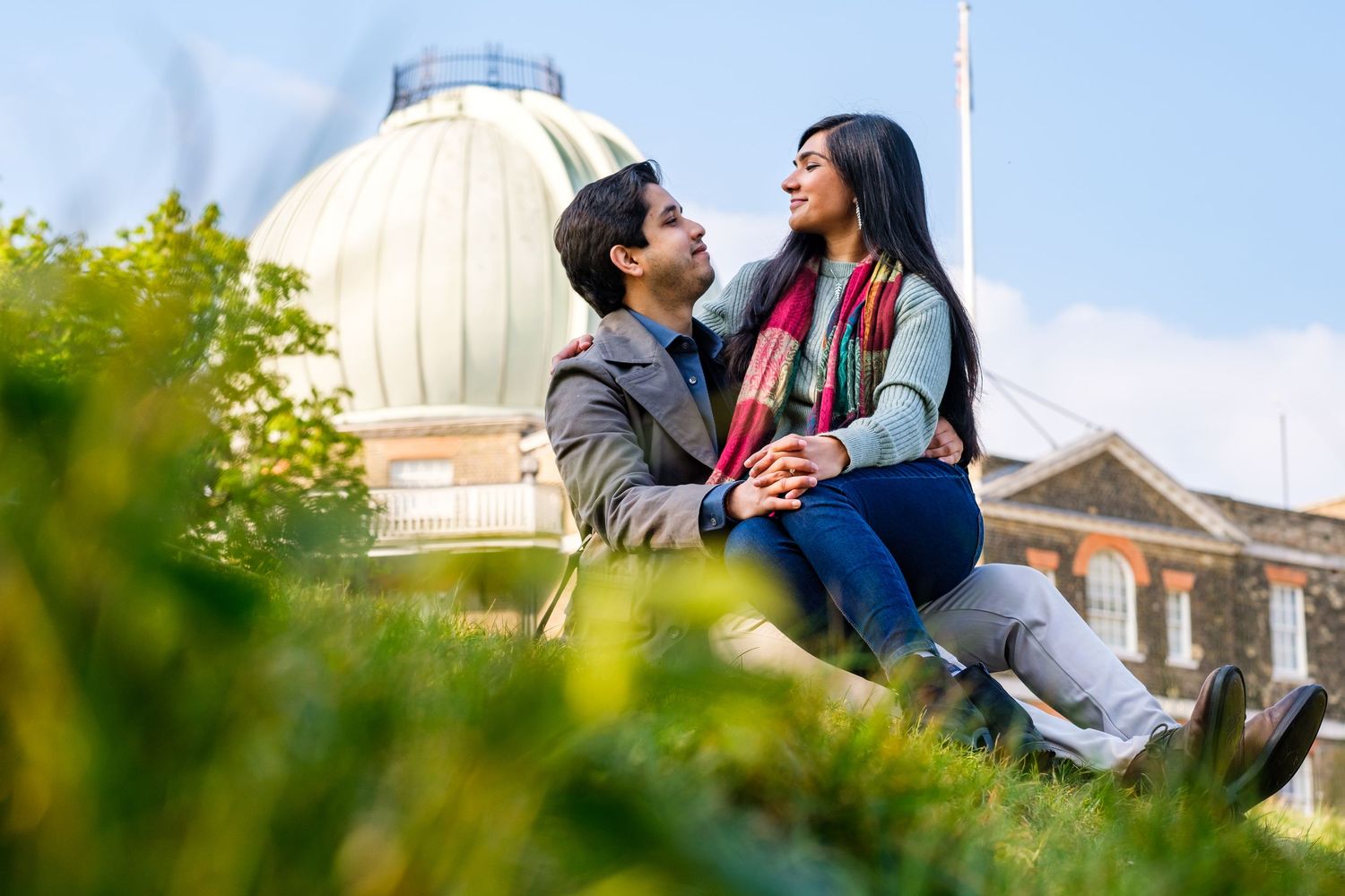 A couple sits together on grass enjoying a sunny day at a university campus with architectural buildings in background.