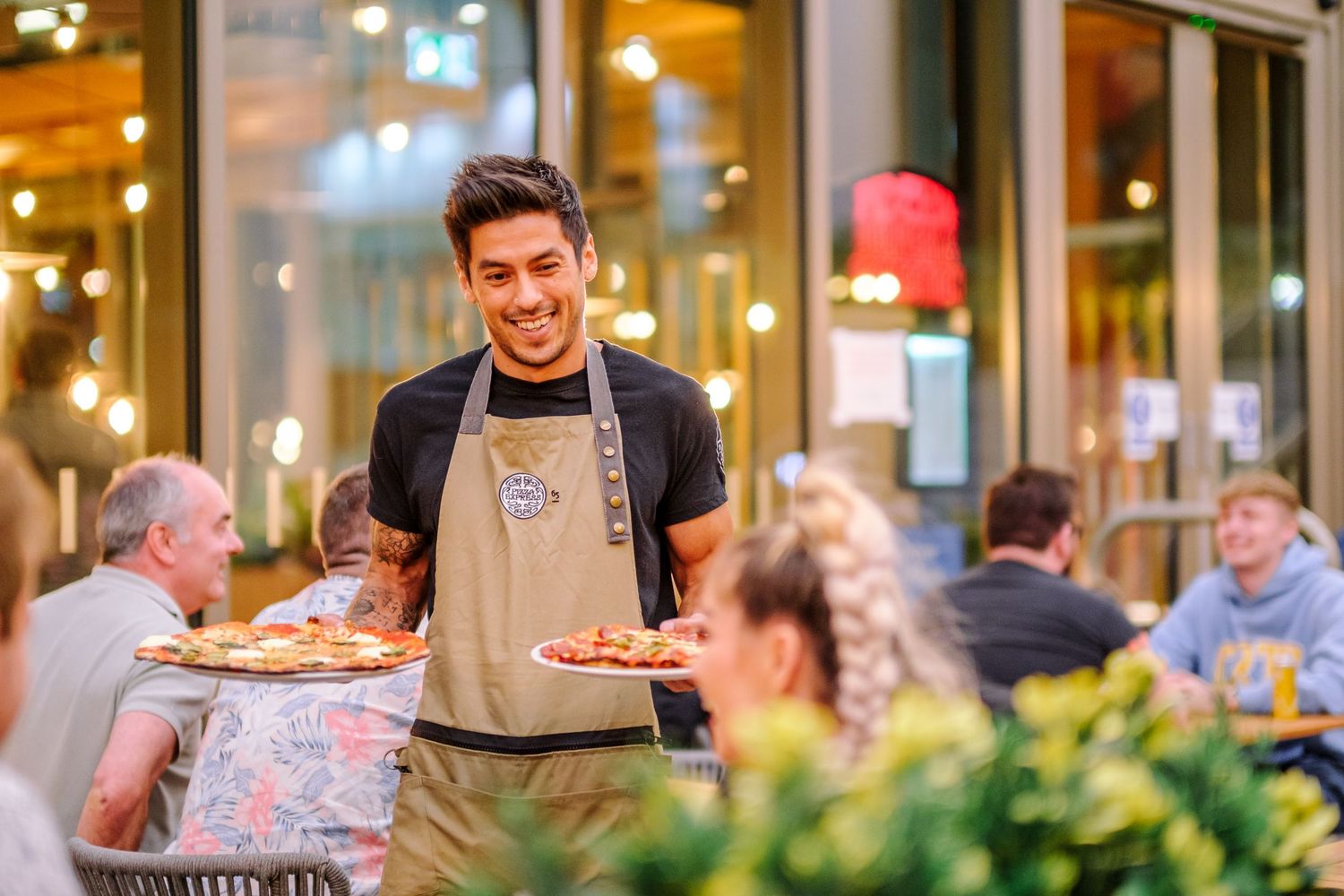 Restaurant server carries plates of food through an elegant dining room with warm lighting and outdoor patio seating.