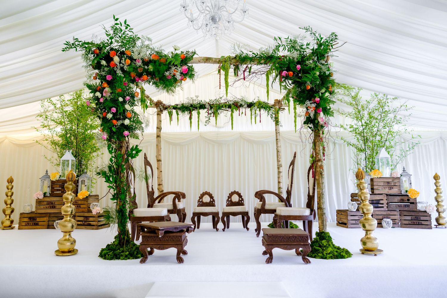 Elegant wedding mandap decorated with greenery and red flowers under a white tent with wooden chairs.