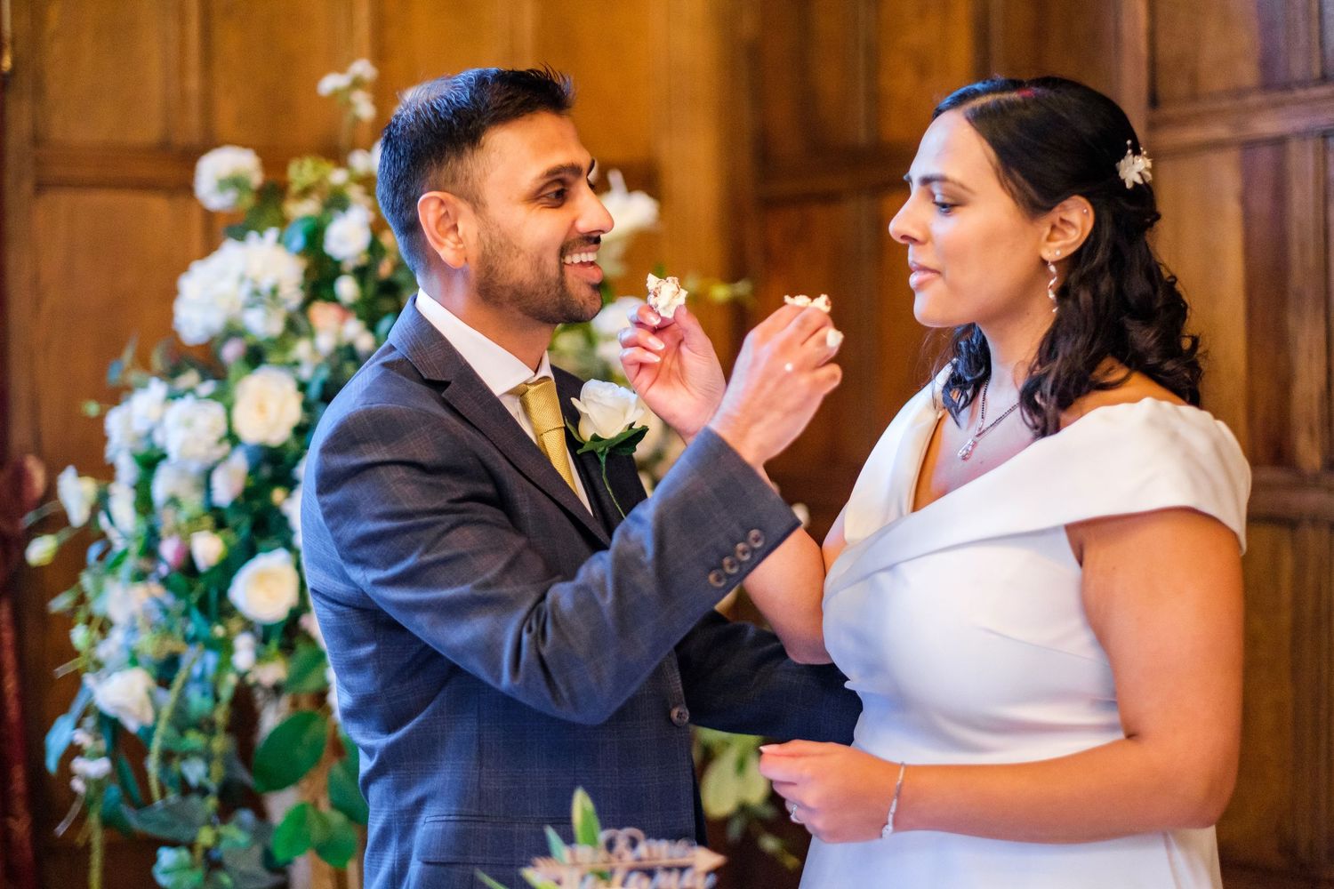 Newlyweds share their first bite of wedding cake together in front of elegant floral arrangements at their reception.