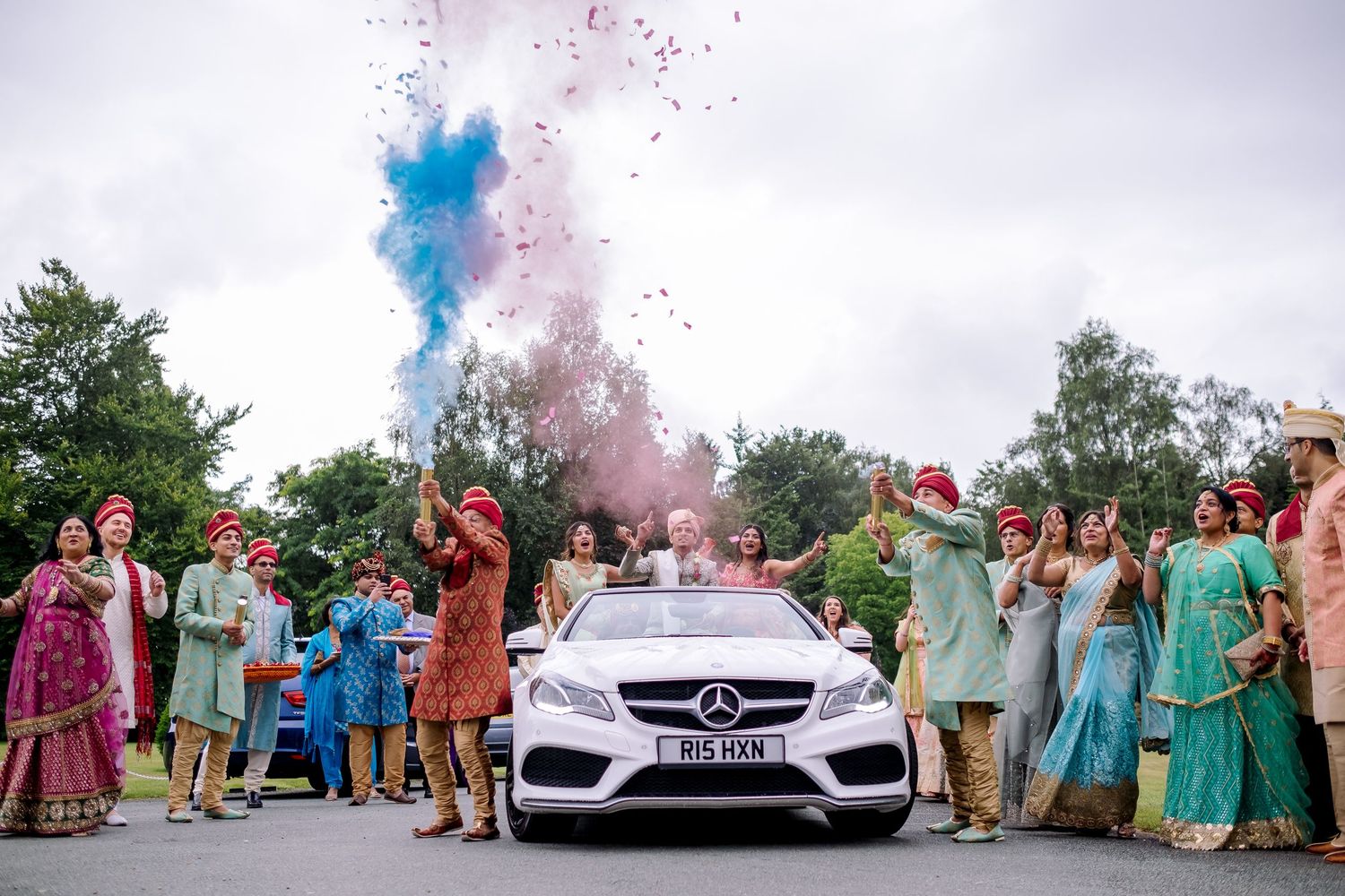 Festive wedding celebration with pink and blue smoke bombs in front of a luxury car.