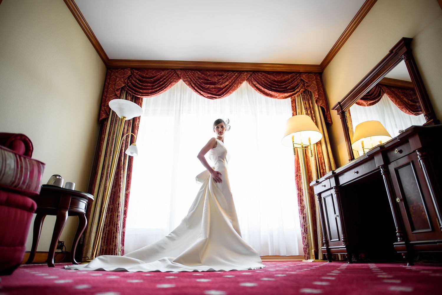 Wedding dress displayed in ornate room with dramatic red curtains and elegant furnishings.