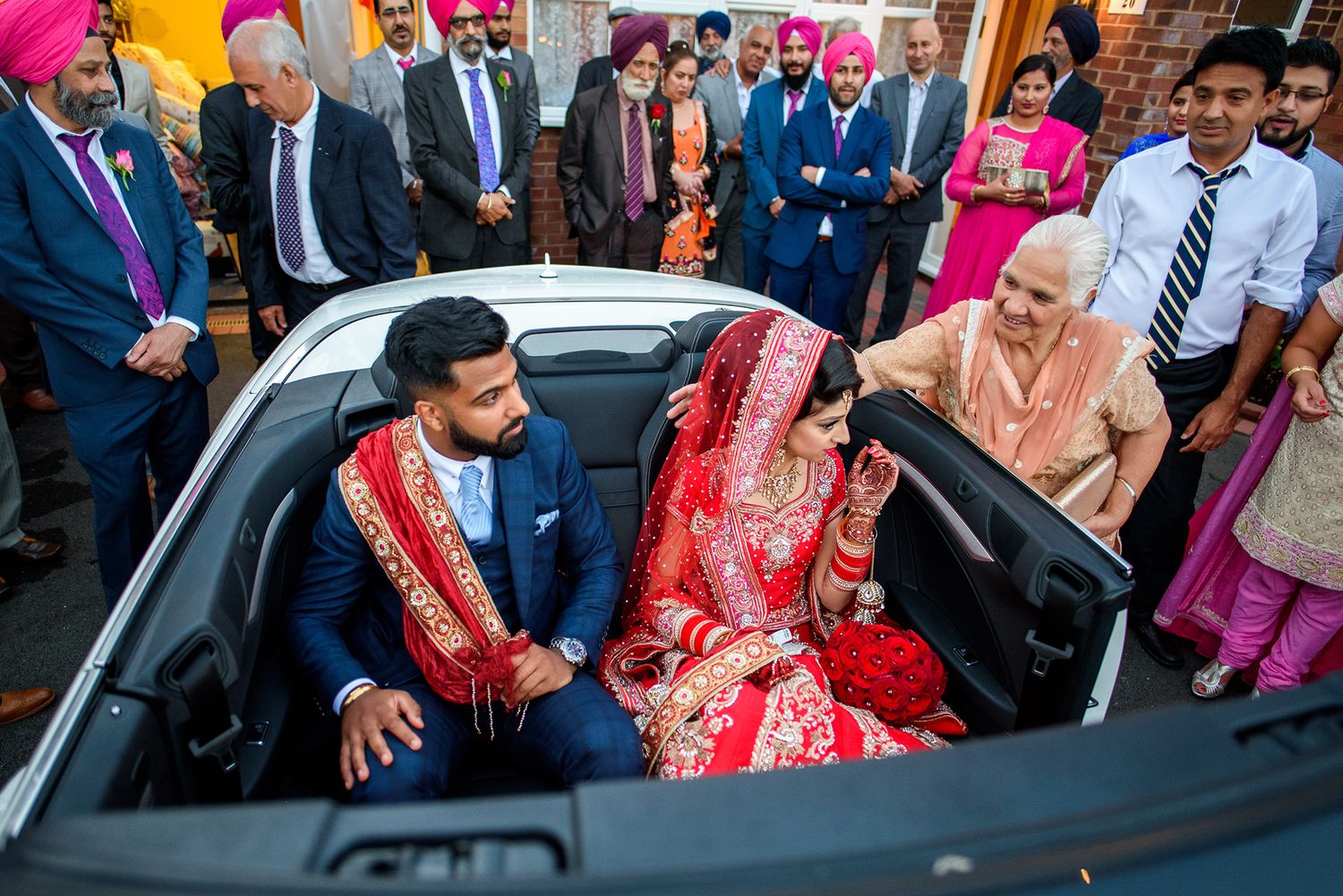 Wedding party and guests celebrating outside with luxury vehicles in formal attire.