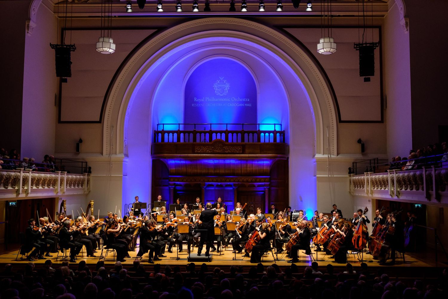 Orchestra performing in an elegant concert hall with arched architecture and blue lighting.