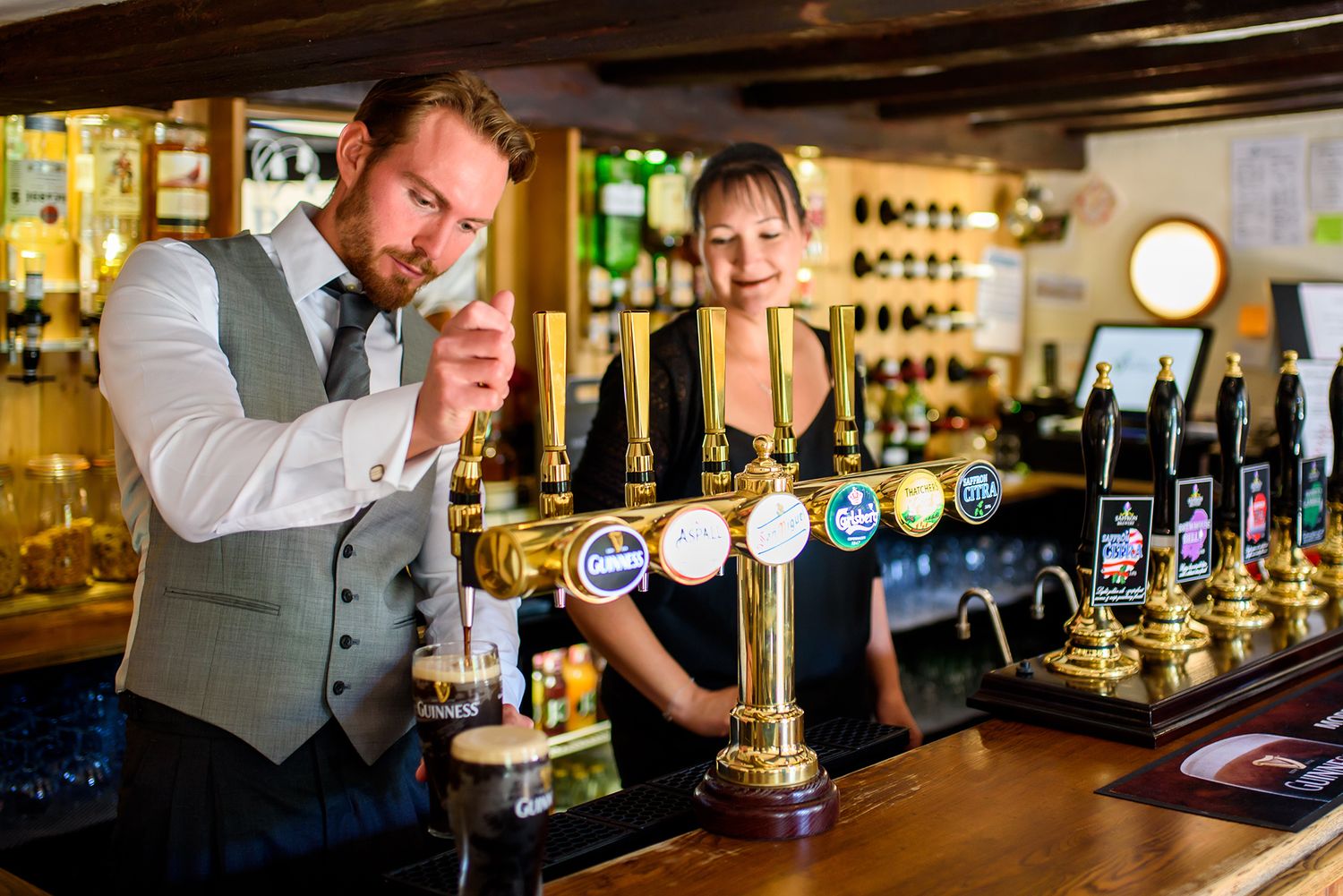Professional bartenders preparing drinks behind a well-stocked wooden bar counter with bottles and taps.