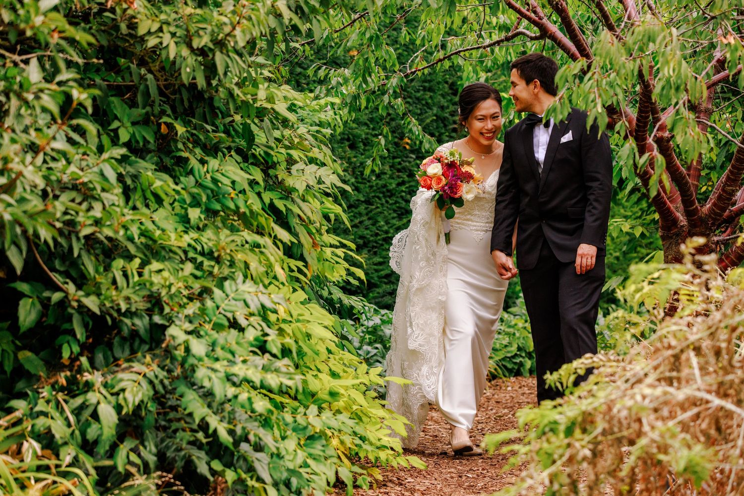 A couple in wedding attire walks along a lush garden path surrounded by greenery.