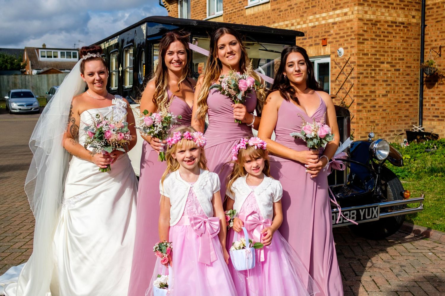 Wedding party members in pink bridesmaid dresses pose together outside a brick building on a sunny day.