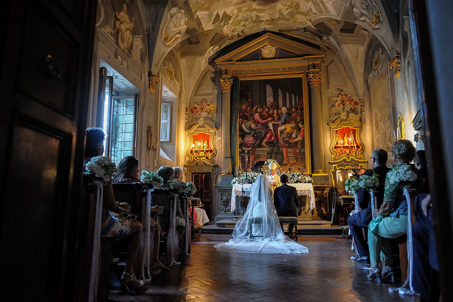 Wedding ceremony in a dimly lit historic chapel with dramatic lighting and frescoed walls.