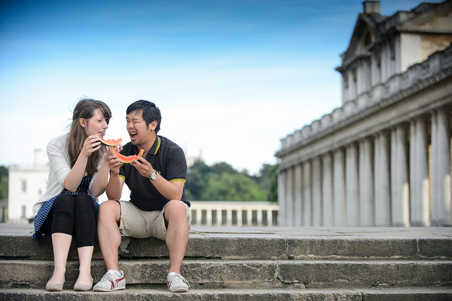 A couple shares a snack while sitting on stone steps in front of a classical building with columns.
