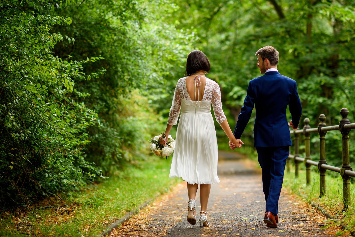 Couple walking along a lush green garden path surrounded by trees and foliage.