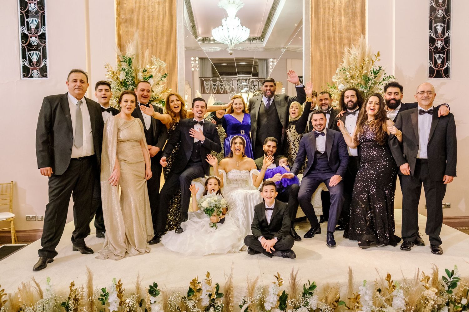 A large group poses together at a formal event with floral decorations in an ornate venue.