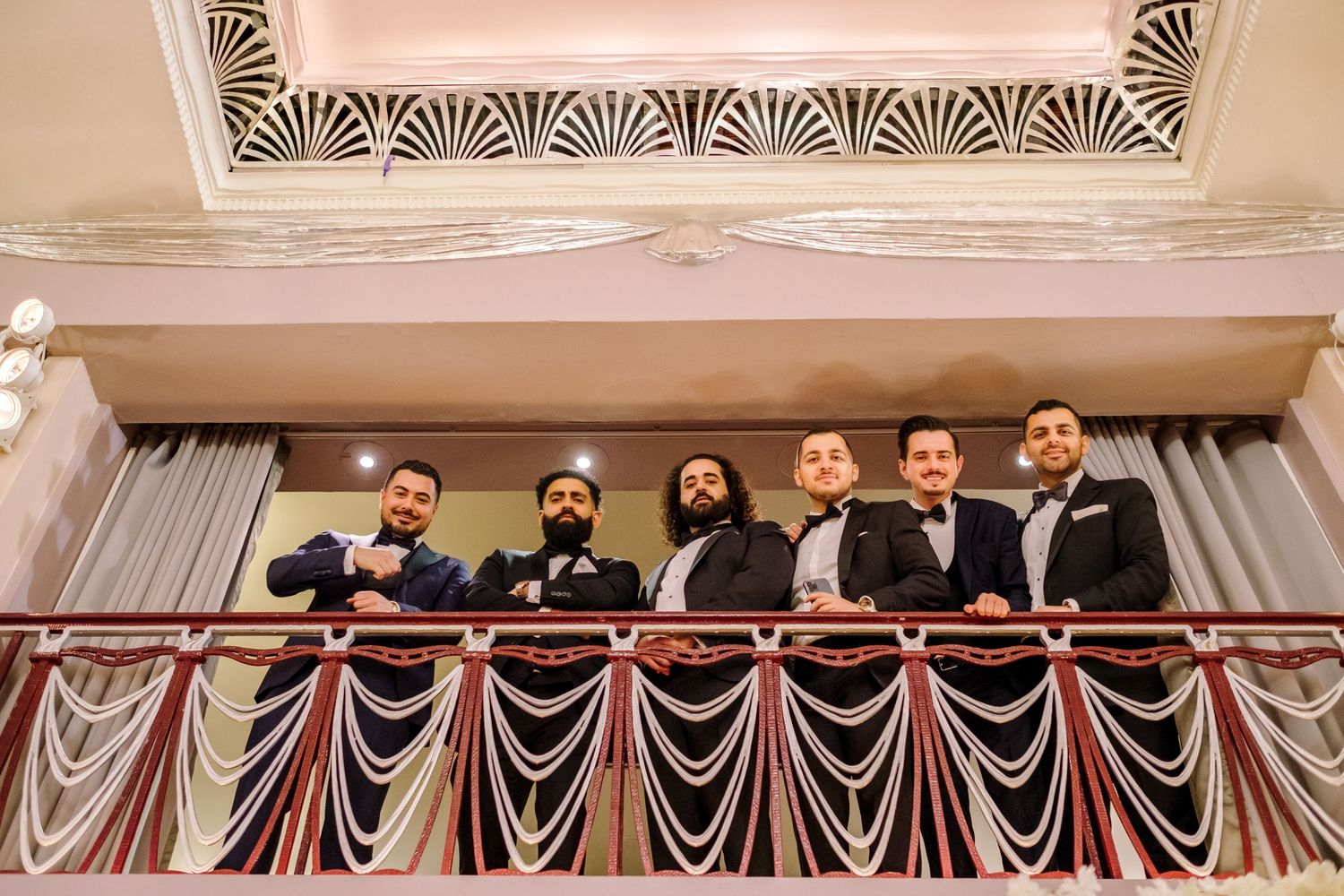 Wedding party members stand in a row on a balcony decorated with draped fabric during an elegant celebration.
