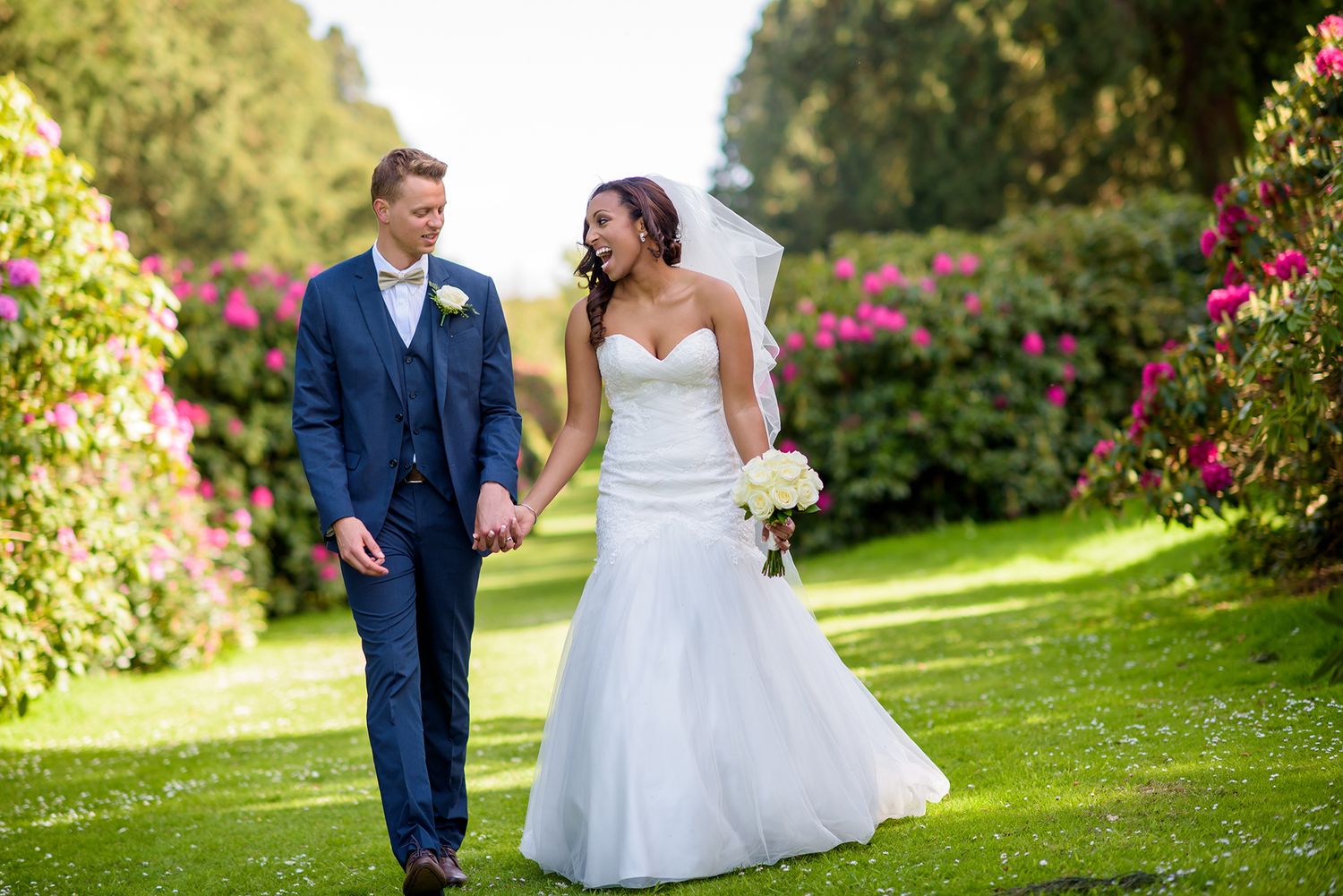 Newlyweds pose together on manicured lawn with blooming flowers.