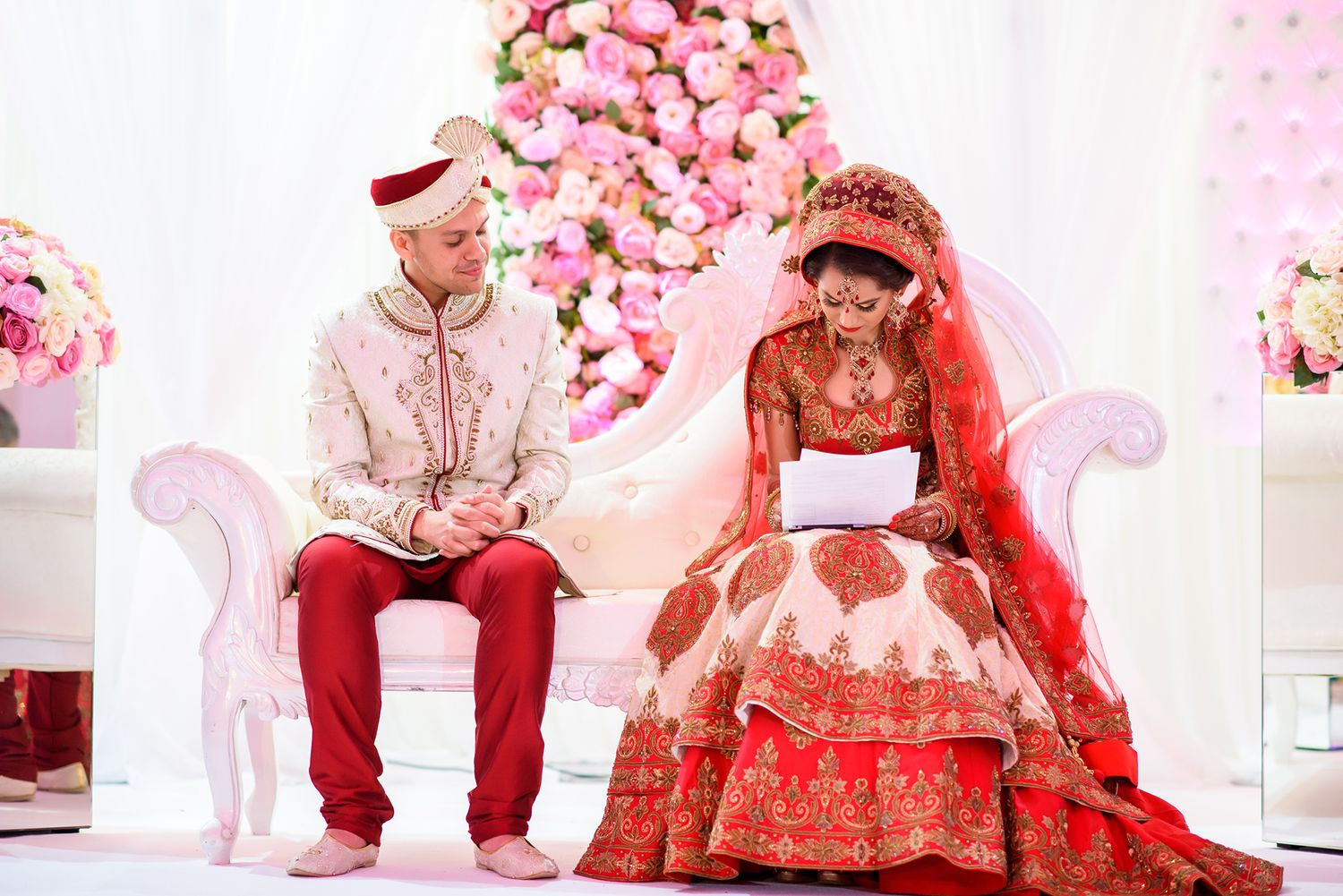 Couple seated on ornate chairs with pink floral backdrop during wedding ceremony.