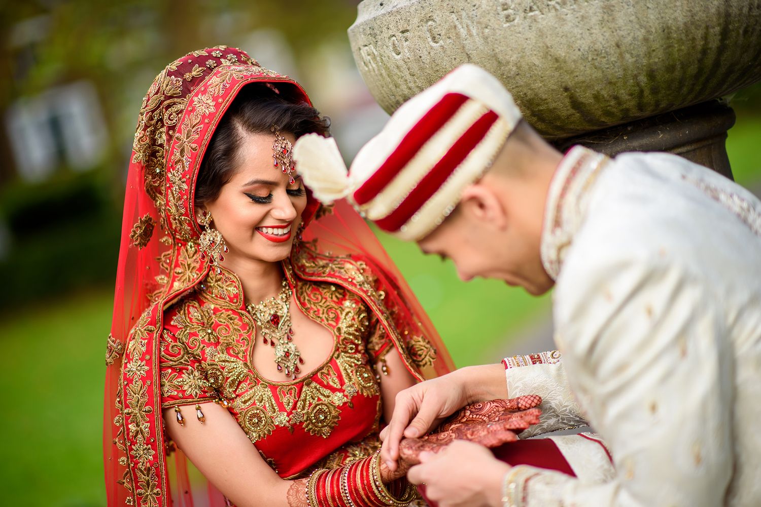 A traditional Indian wedding couple in red and white attire pose together in an outdoor garden setting.