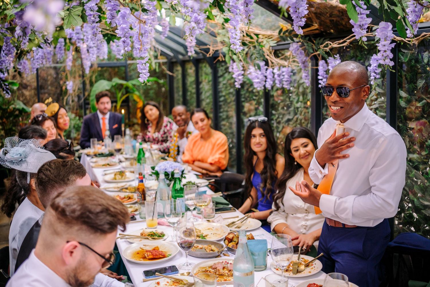 Wedding reception guests enjoying an outdoor dinner under wisteria and garden lights.