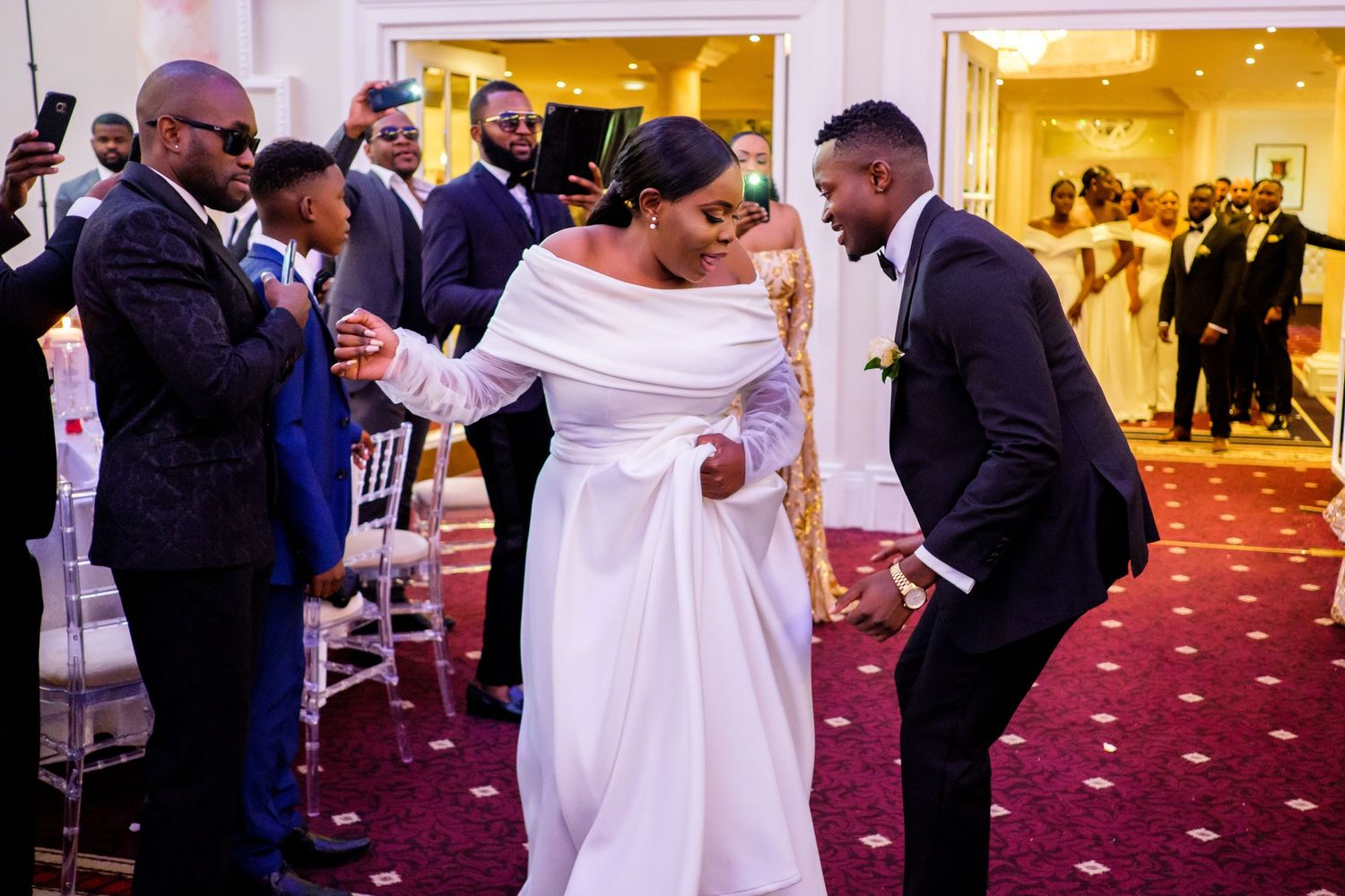 Guests dance and socialize during a wedding reception in a luxurious ballroom with red carpeting.