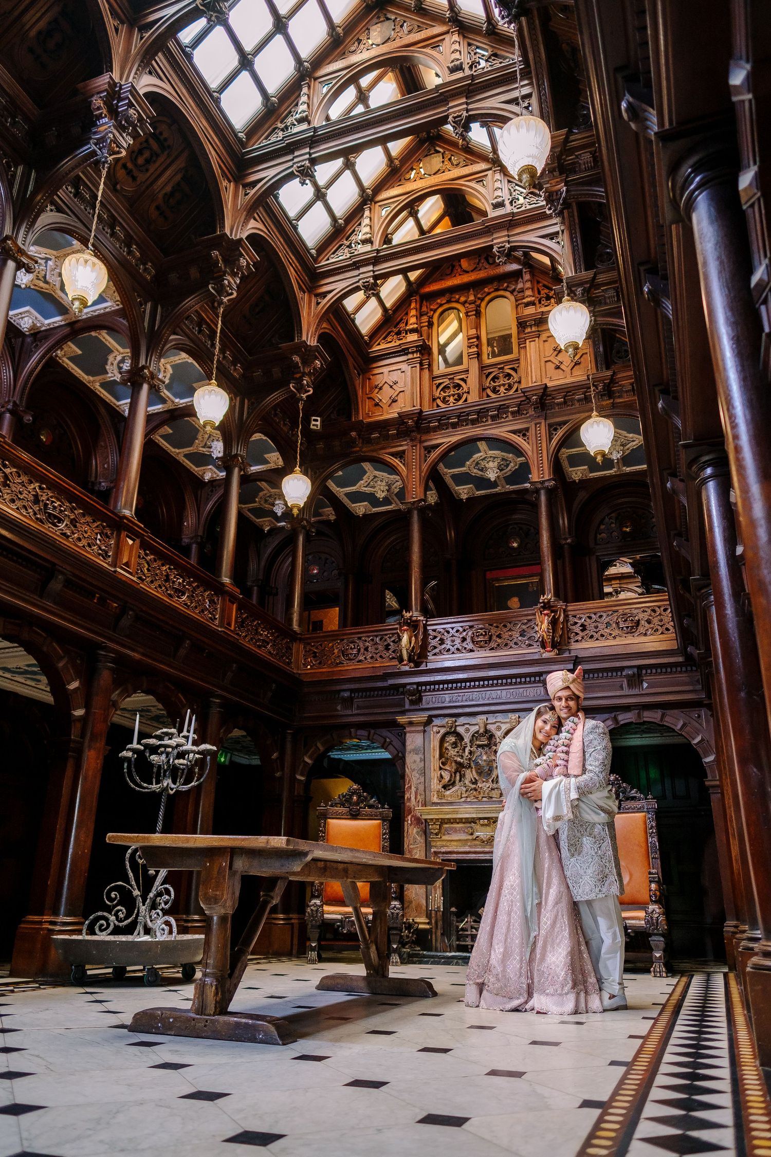 Couple in wedding attire poses in ornate Victorian gothic interior with wooden panels and hanging lamps.