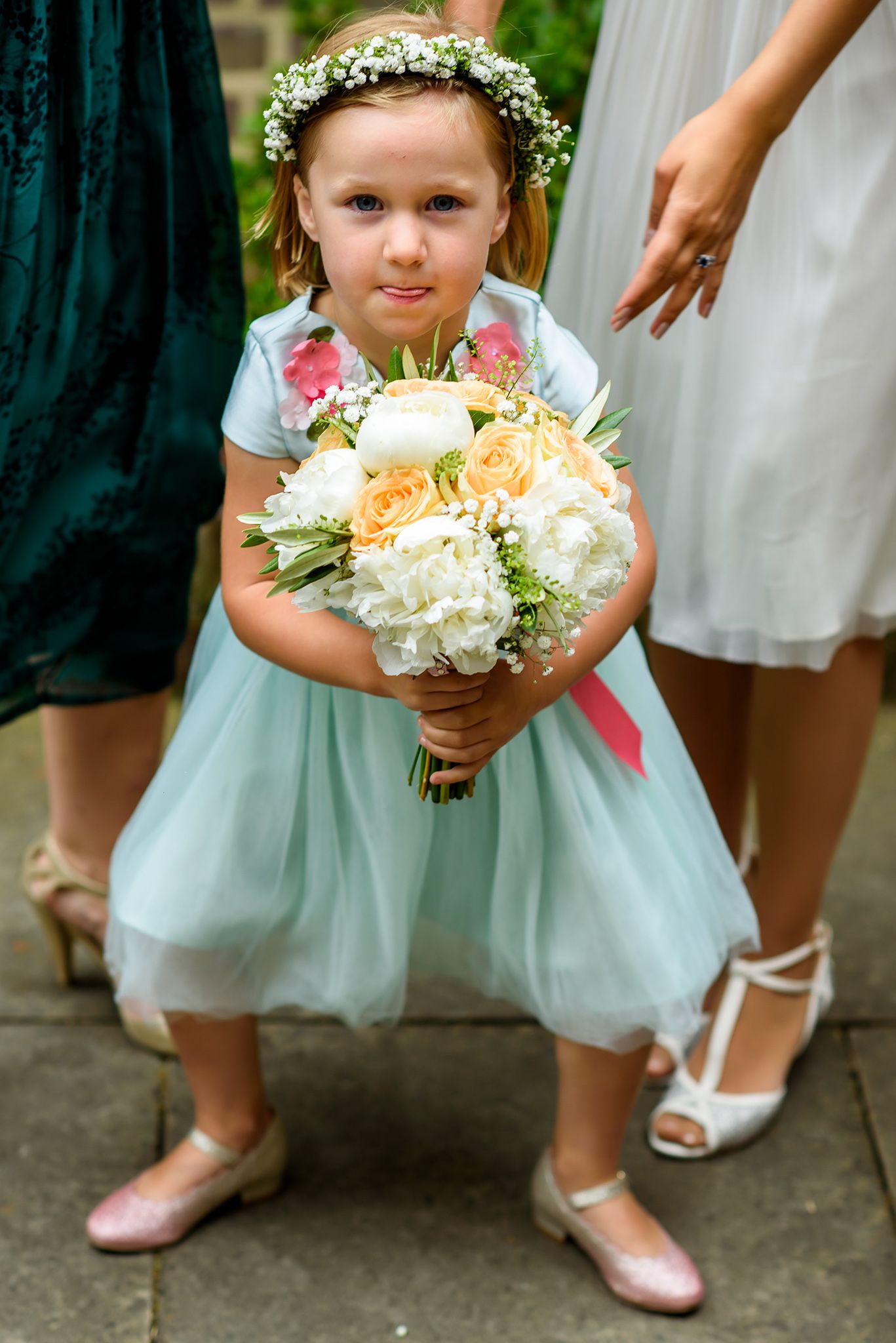 A young flower girl in a mint dress holds a peach and white bouquet while wearing a flower crown.