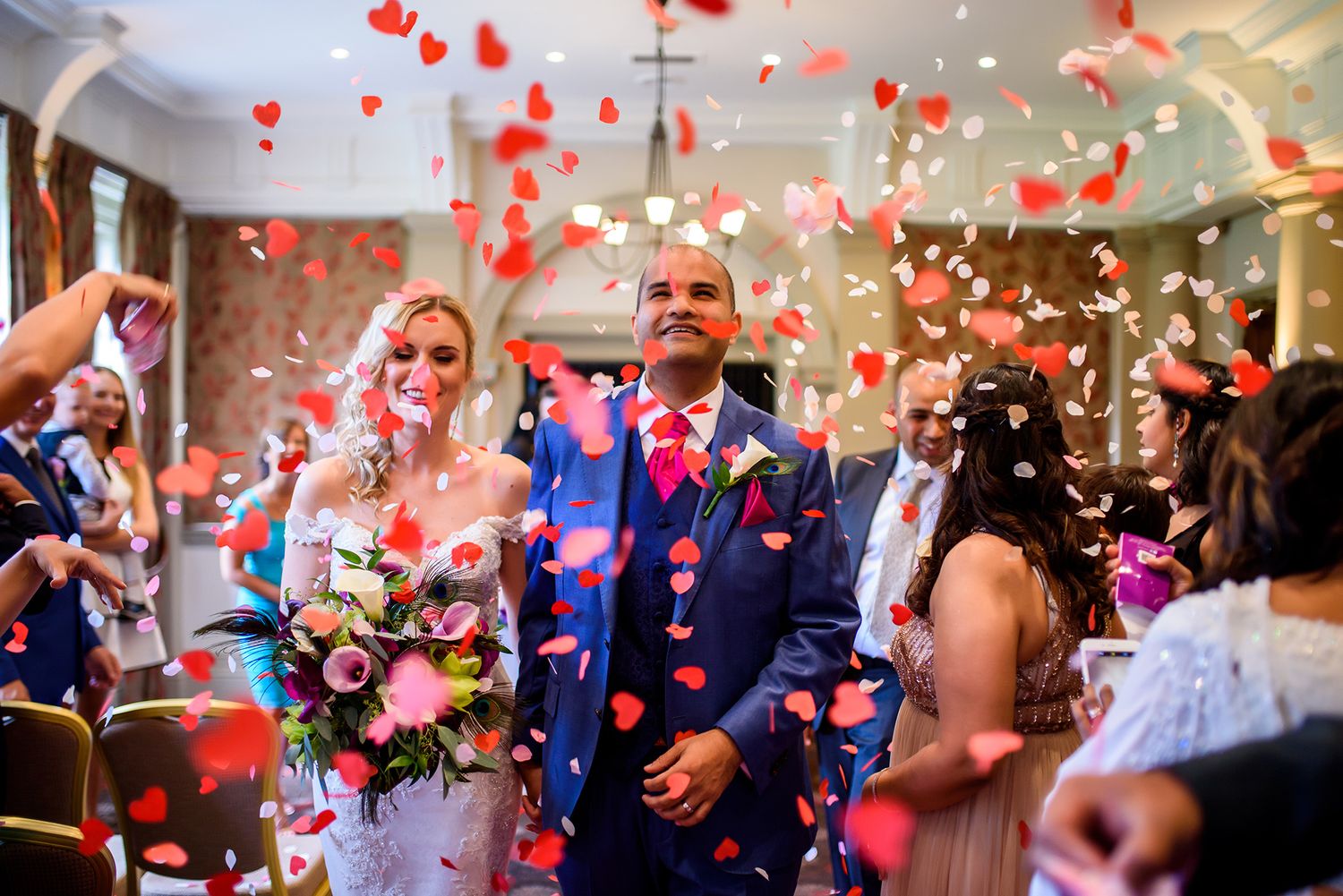 Wedding party celebrates under a shower of red heart-shaped confetti in an elegant venue.