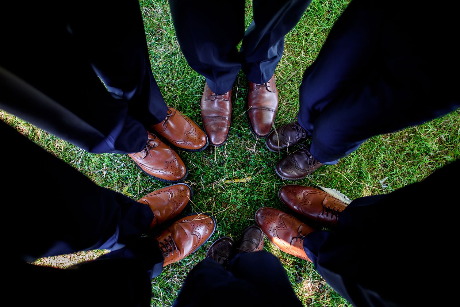 A circle of dress shoes and suit pants photographed from above on green grass.