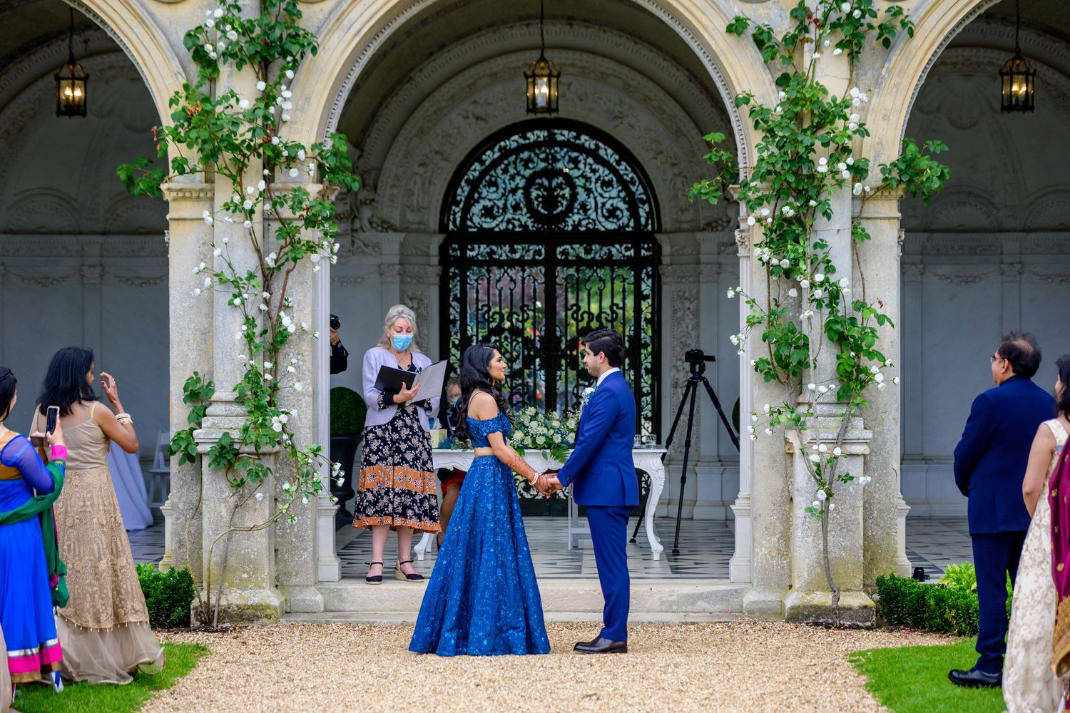 Wedding guests gather under elegant stone archways decorated with climbing vines at a stately venue.