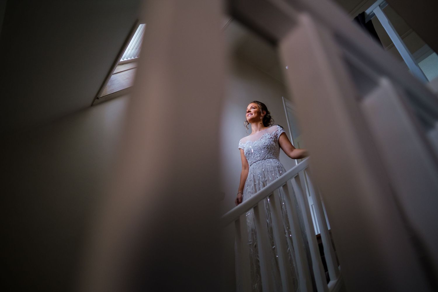 A bride in a white dress poses dramatically on a staircase with natural window lighting creating an artistic effect.
