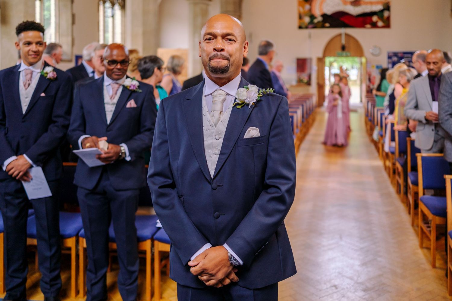 A groom stands expectantly at the front of a traditional church aisle during a wedding ceremony.
