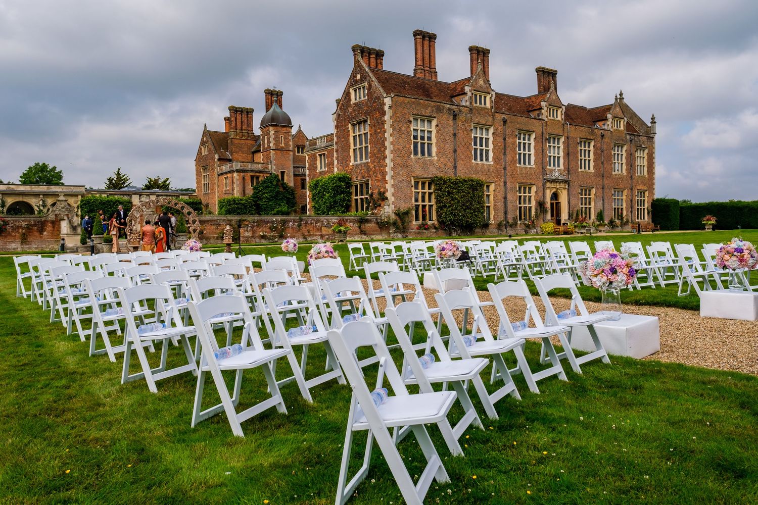 White chairs arranged on a lawn for an outdoor wedding ceremony in front of a grand historic brick mansion.