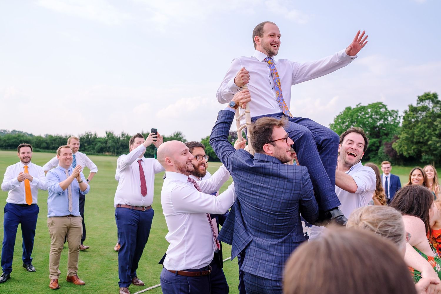 Wedding party members playfully posing and celebrating on sunny outdoor lawn.