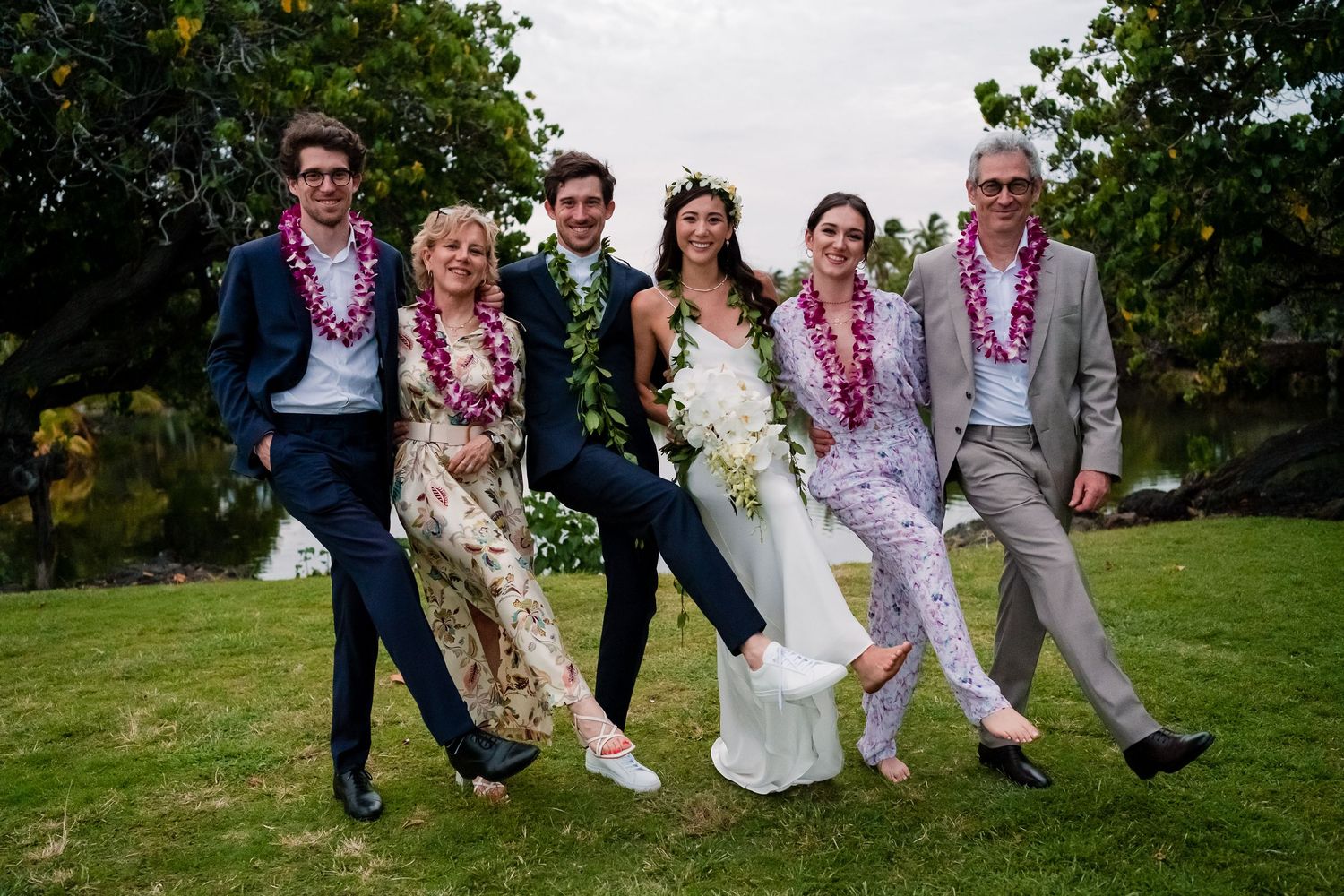 Group photo of wedding party sitting together on grass with leis and flowers in outdoor garden setting.