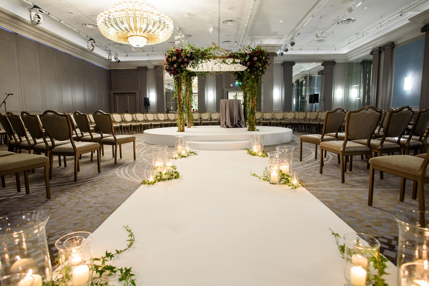 Luxurious wedding ceremony setup with white aisle runner, floral arch, and crystal chandelier in an elegant ballroom.