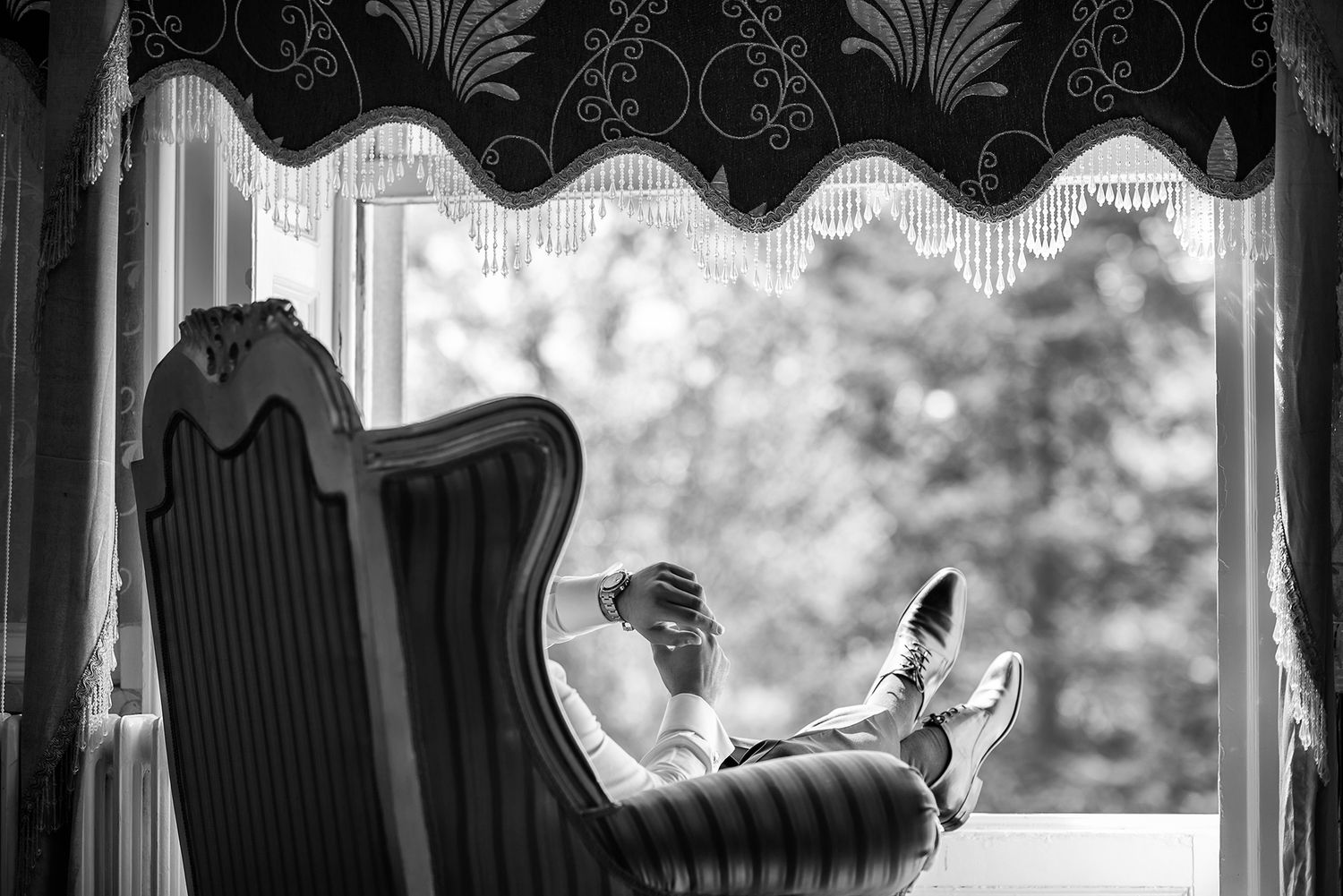 A relaxed figure sits with feet up in an elegant vintage chair by a window with decorative curtains.