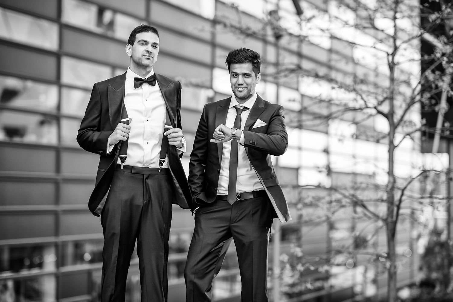 Two men in formal attire pose outdoors near a modern building.