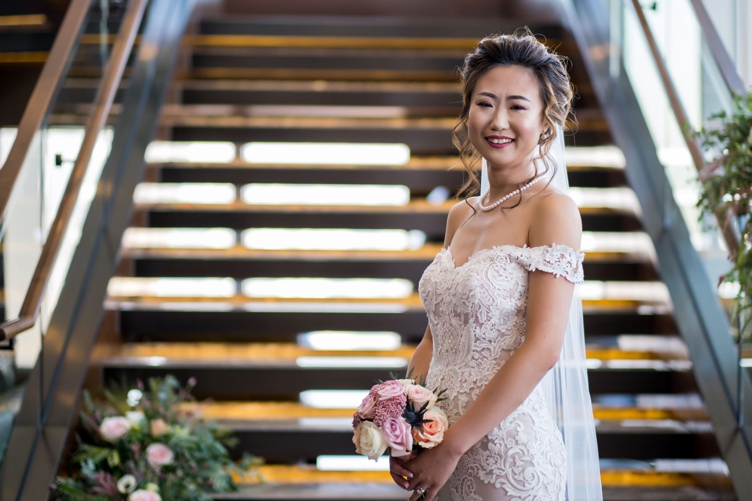 A bride in a lace off-shoulder wedding gown poses elegantly in a modern architectural space with wooden beams.