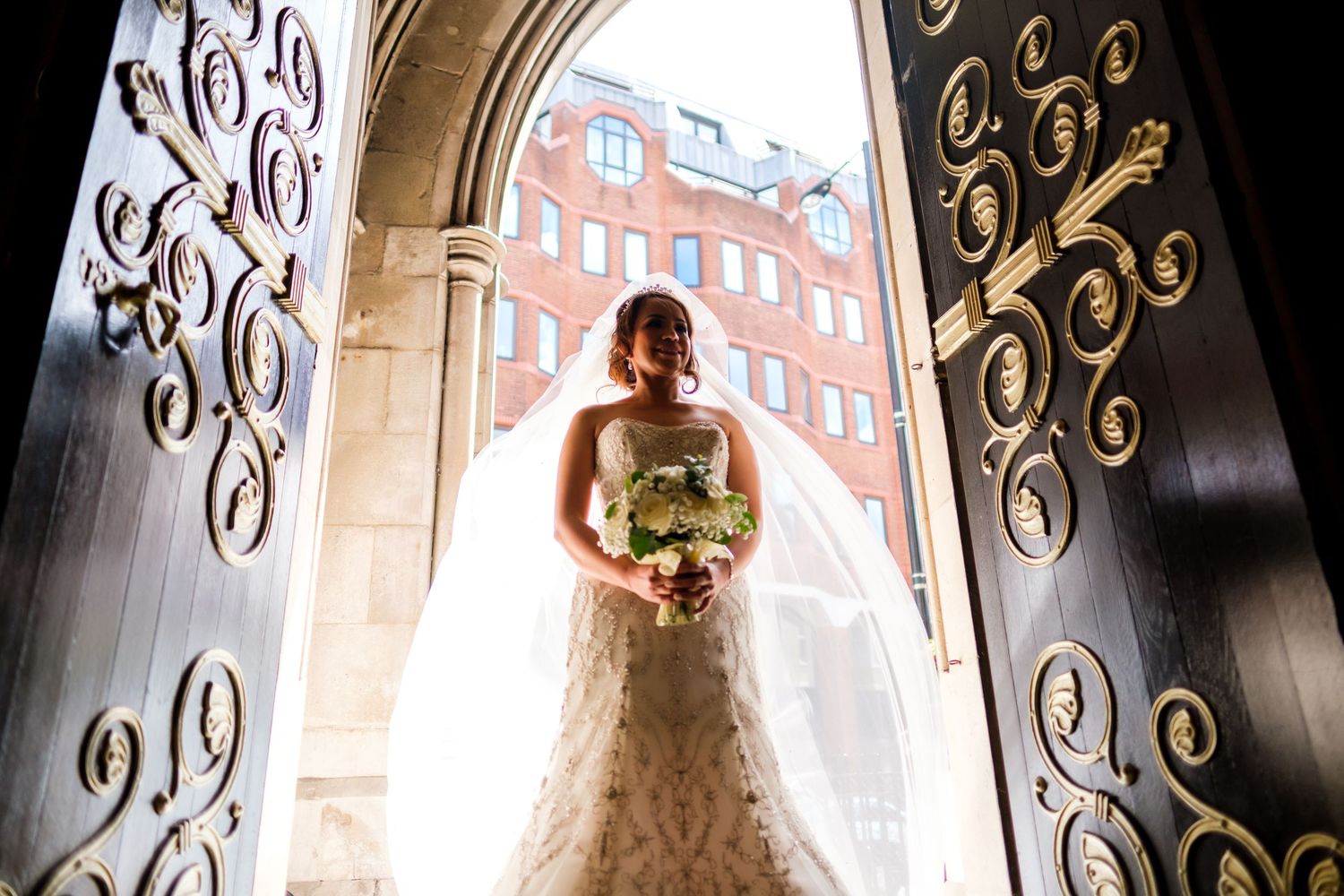 A bride in a white wedding gown stands in an ornate doorway with wrought iron details and brick architecture.