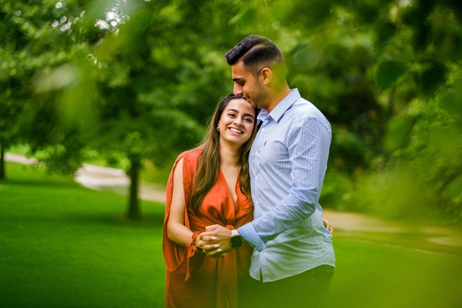 A couple sharing a romantic embrace while walking along a tree-lined path in a verdant park.