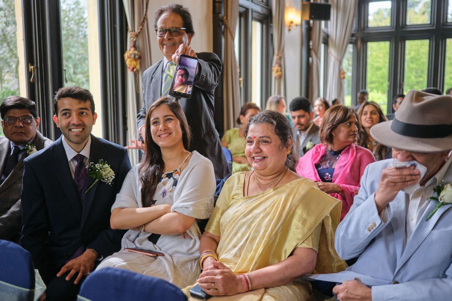 Wedding guests smile and laugh while seated in a bright room with large windows during a celebration.