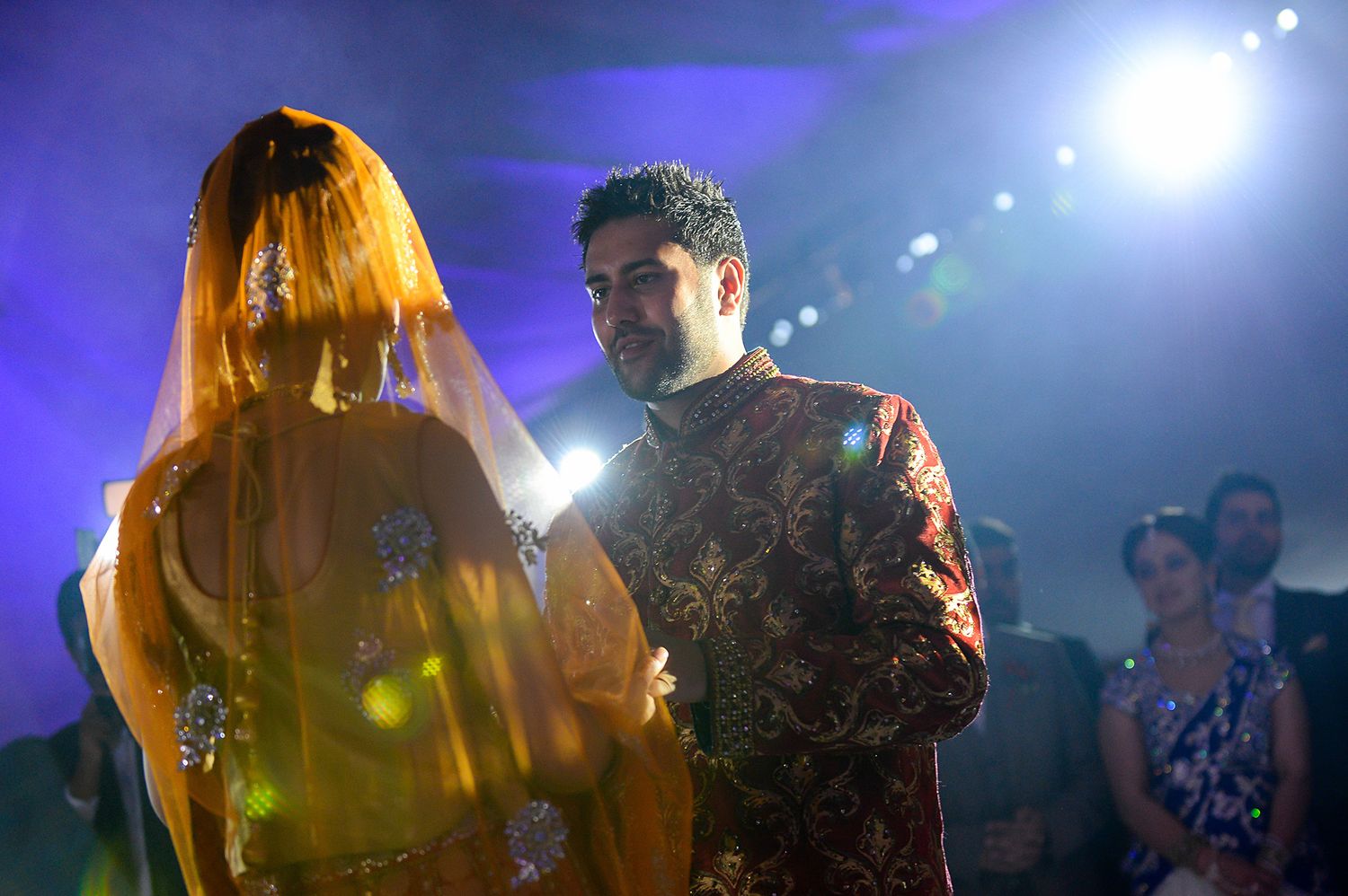 Bride in yellow veil stands with groom during traditional ceremony under blue lighting.