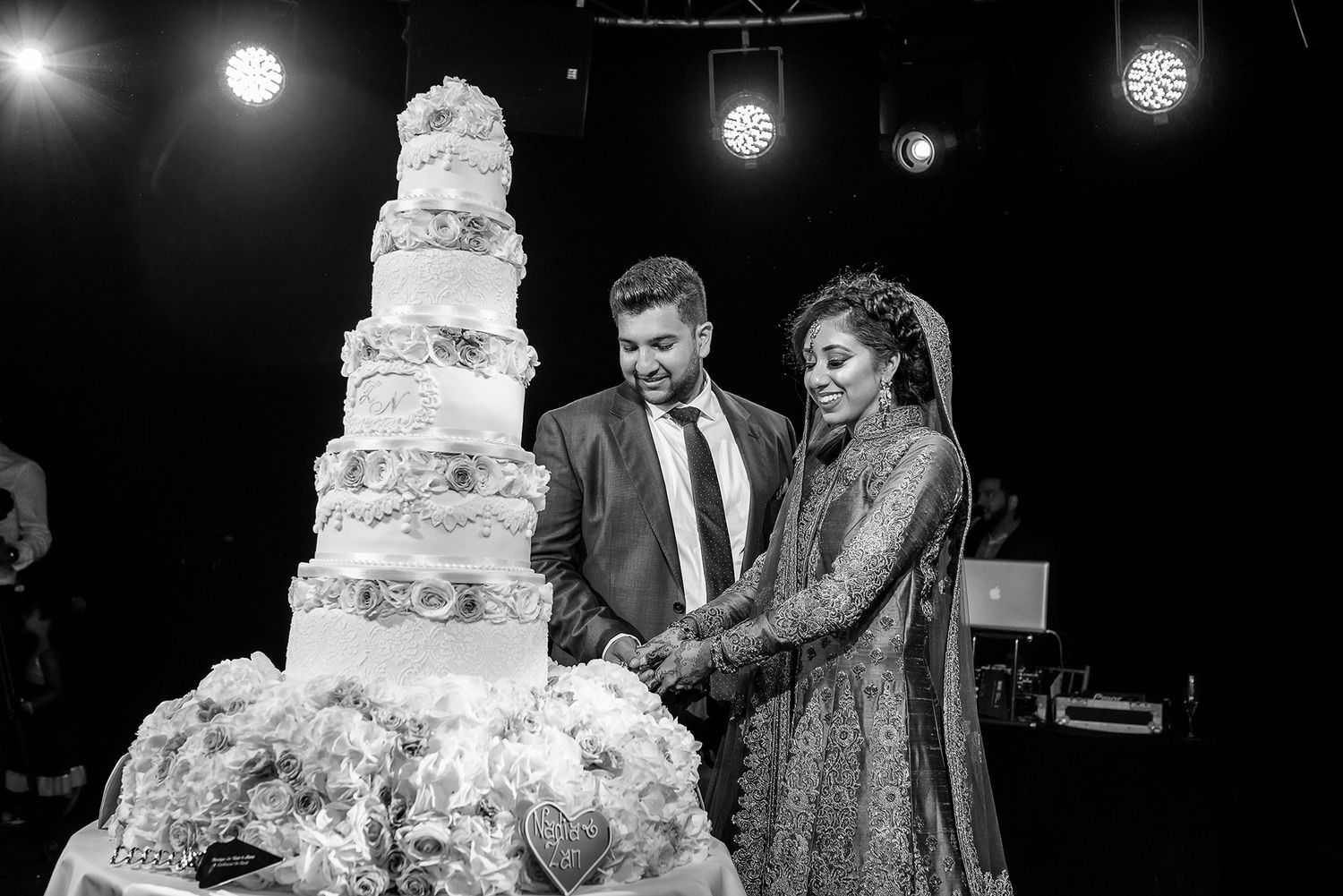 Couple cutting an elaborate multi-tiered white wedding cake under hanging lights.