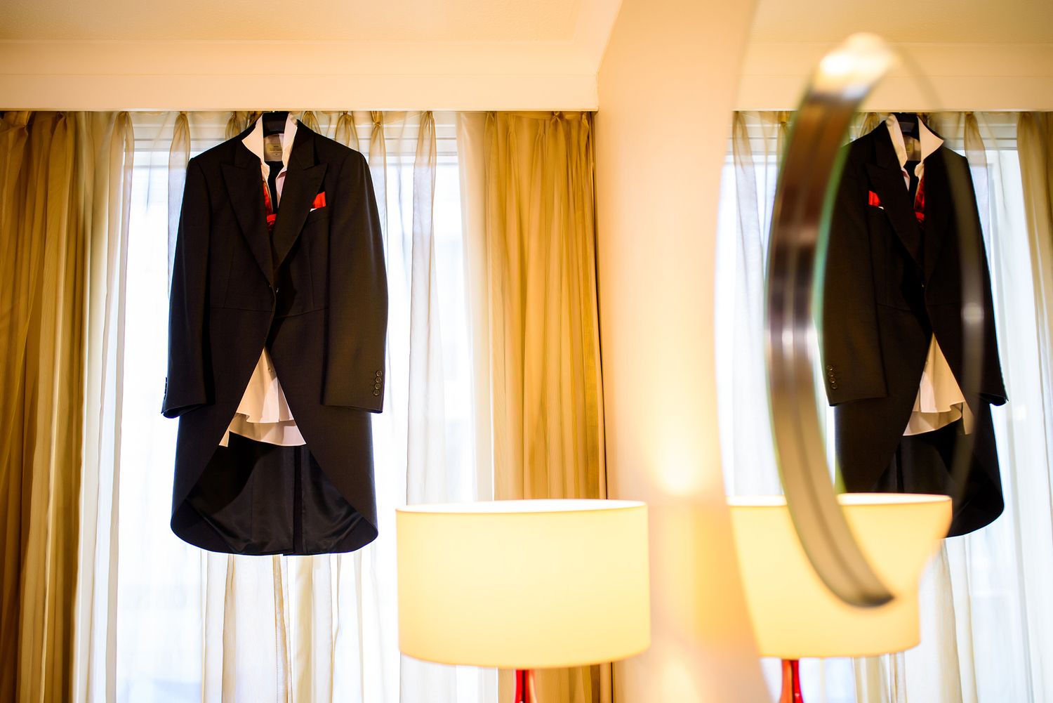 Wedding suits hanging by window with warm natural lighting and golden curtains.