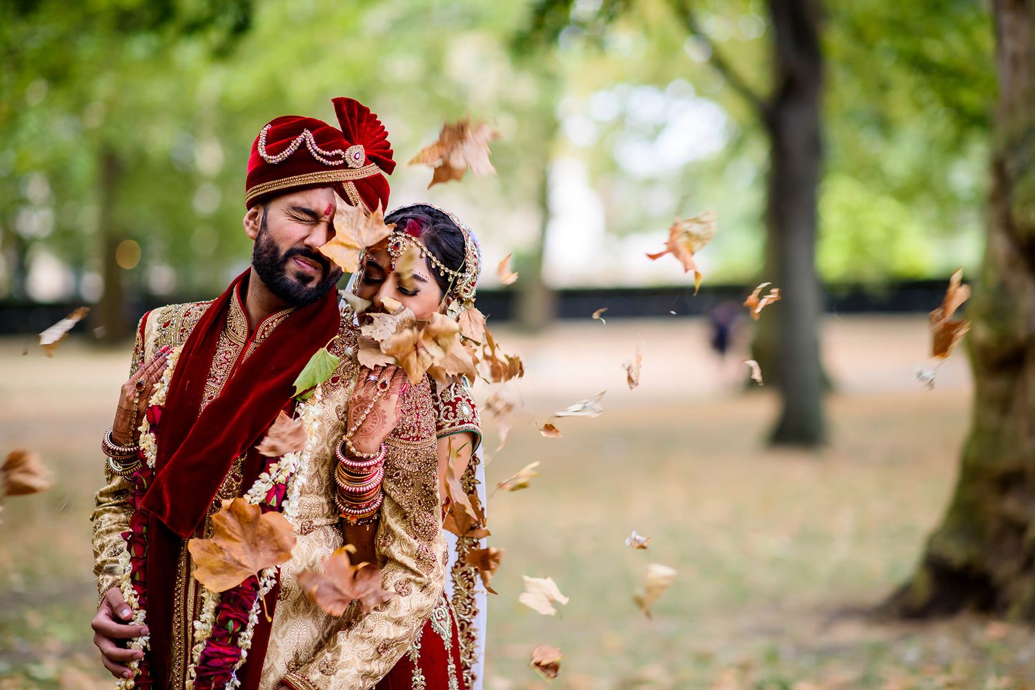 Fall leaves scatter around a couple in traditional Indian wedding attire in a park setting.