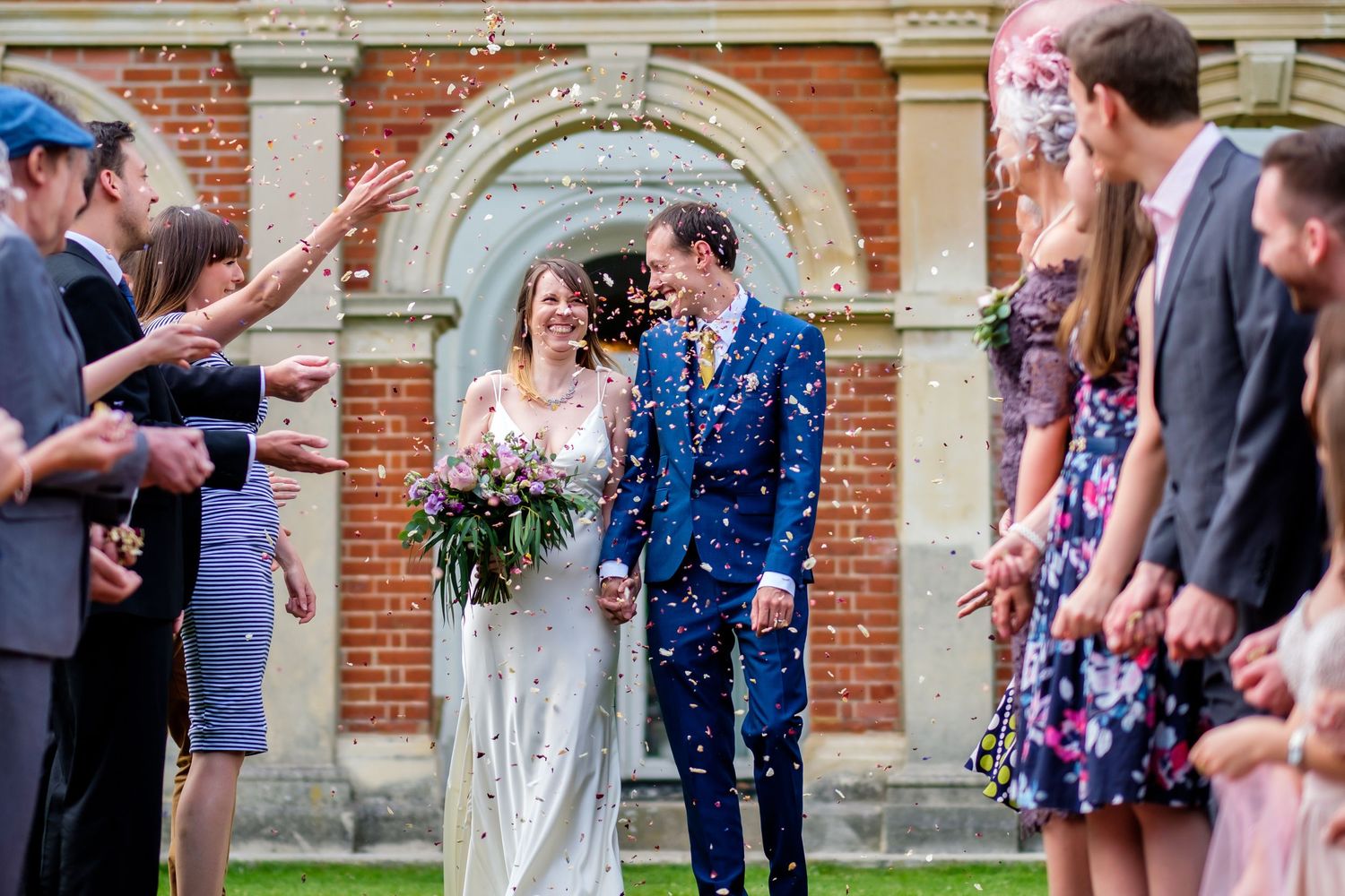 Wedding party and guests celebrate with confetti outside a brick building.