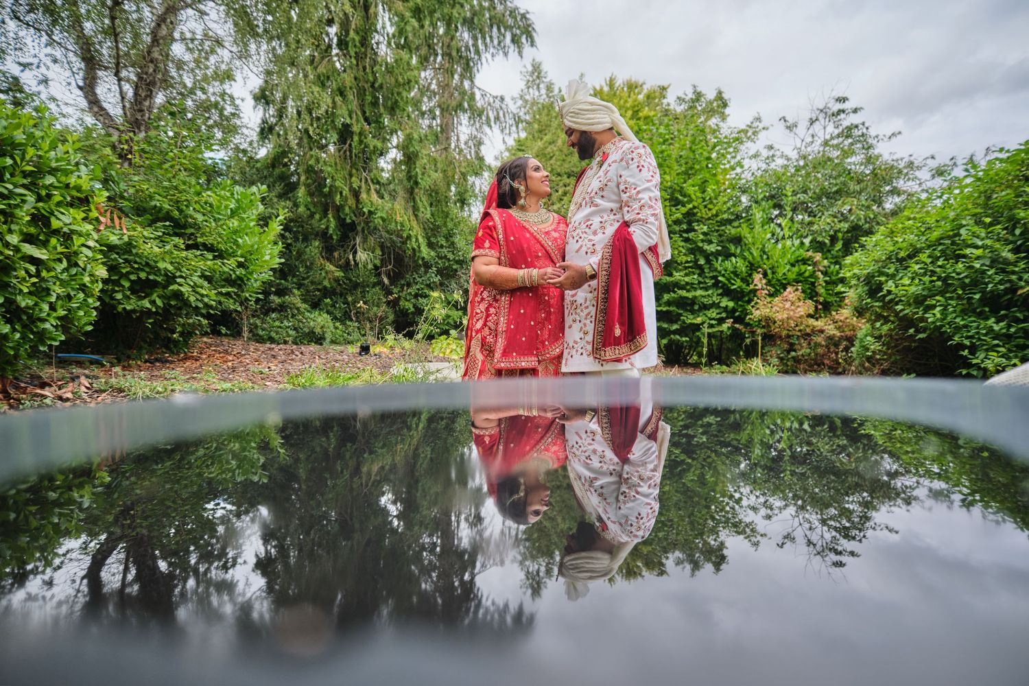 Reflection of people in traditional Indian wedding attire walking along a serene pond with green foliage.