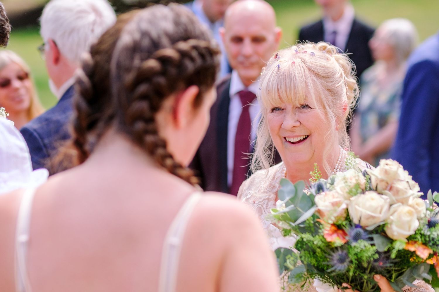 Wedding guests gather outdoors on a sunny day celebrating with joyful expressions and floral bouquets.