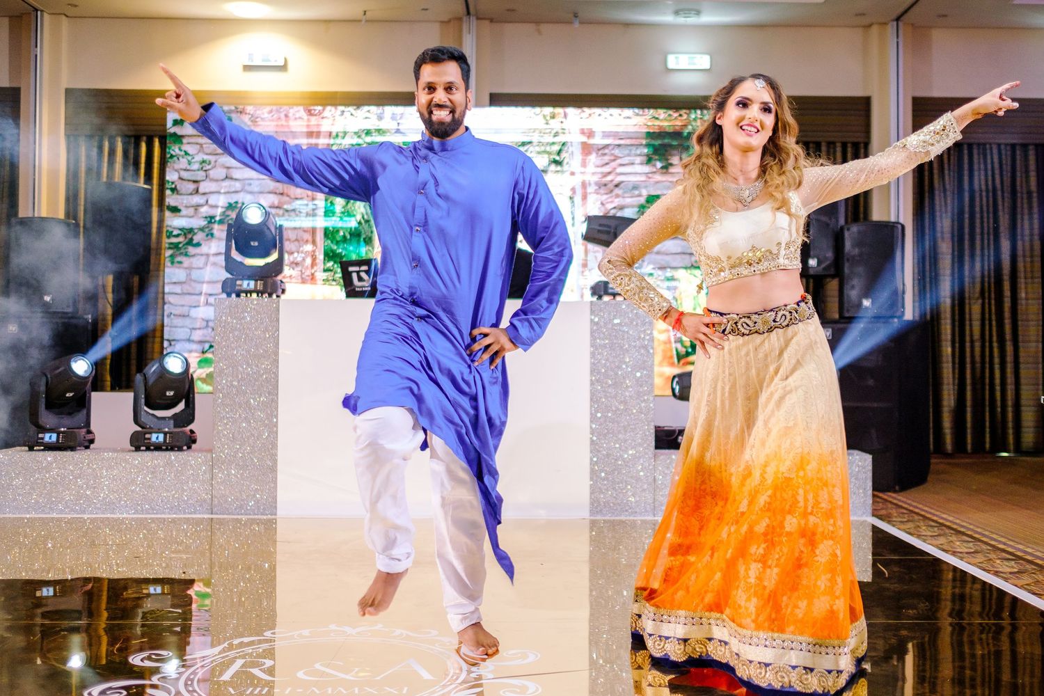 Couple performs energetic dance in fusion Indian wedding attire on indoor stage.
